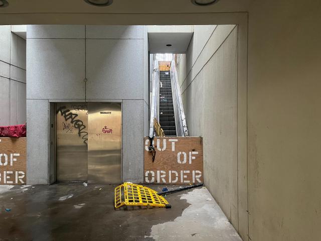 Boarded up escalators with OUT OF ORDER spraypainted on the wood board. Surrounded by rubbish. Graffitied elevator.