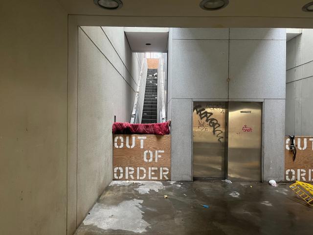 Boarded up escalators with OUT OF ORDER spraypainted on the wood board. Surrounded by rubbish. Graffitied elevator.