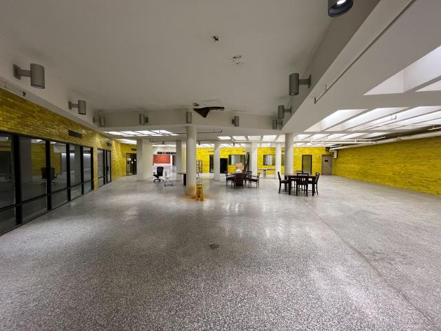 A dark and dingy room with columns supporting the roof. Half working fluorescent lights, leaking ceiling, lino floor. A selection of chairs and tables, and a solitary desk chair with no desk.
