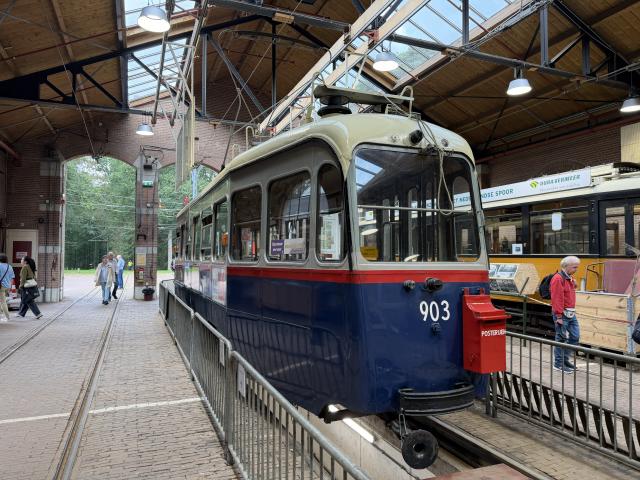 A white, red and blue old tram inside a carriage shed, with a space to walk underneath the tram. It has a post box on the front of it. There are tourists examining it.