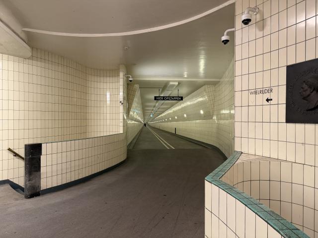 A tiled underground tunnel with a sign reading HIER OPSTAPPEN. Cylists can be seen using it in the distance. It is well lit. There are stairs going down furthur.