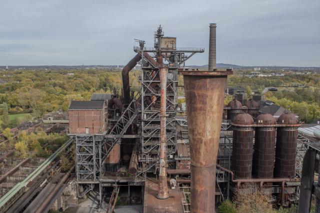 A large, rusted industrial facility with metal structures and a chimney, surrounded by greenery.